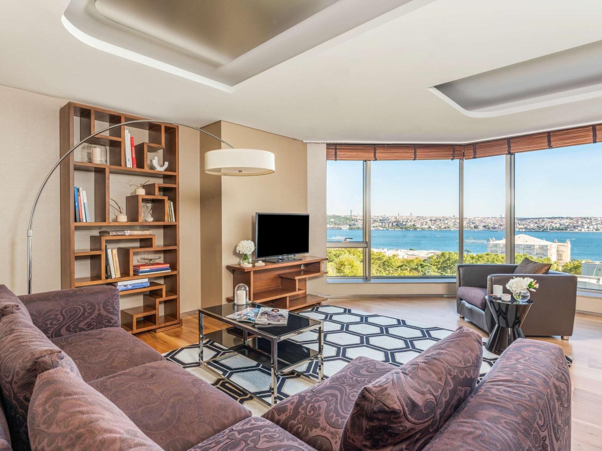 Swissotel The Bosphorus Istanbul Eksteriør billede The photo shows a modern living room with a large window offering a scenic view of a body of water. The room features a brown sectional sofa, a glass coffee table, and a stylish rug with a geometric pattern. There is a contemporary floor lamp and a b