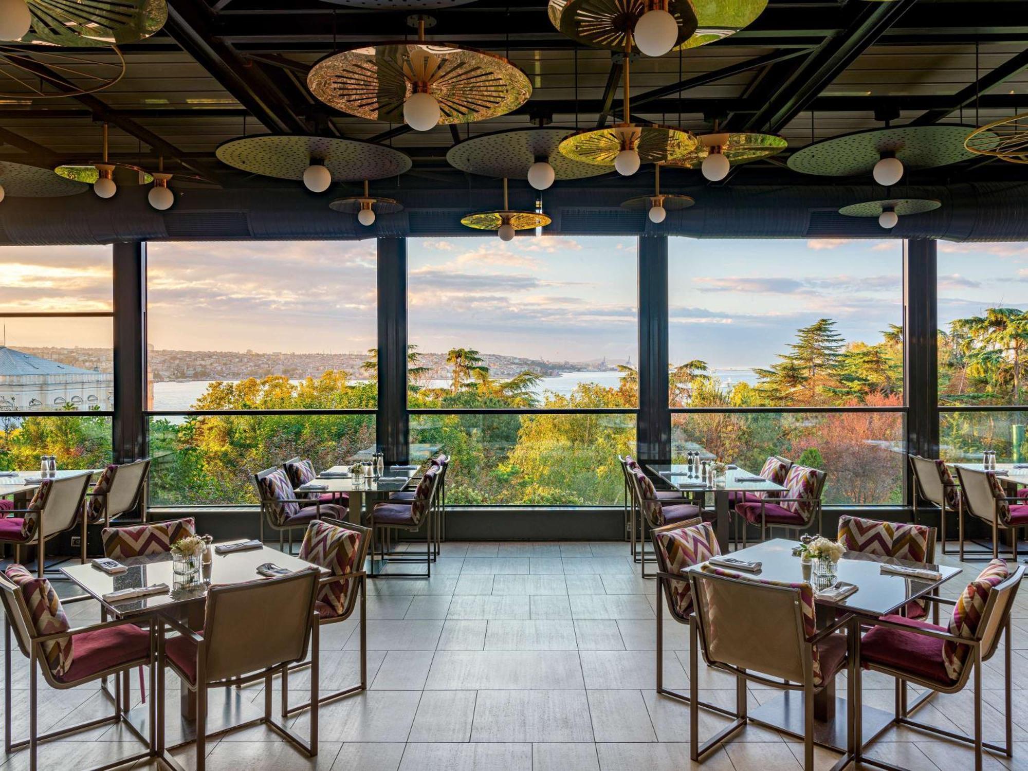 Swissotel The Bosphorus Istanbul Eksteriør billede The photo shows a modern dining area with several tables and chairs arranged neatly. Large floor-to-ceiling windows provide a stunning view of a landscape, likely featuring trees and a body of water. The interior design includes stylish light fixture