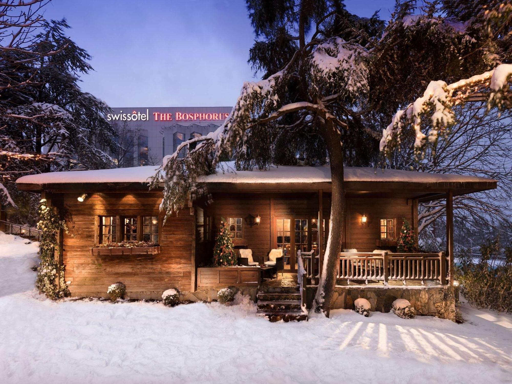 Swissotel The Bosphorus Istanbul Eksteriør billede The photo shows a charming wooden cabin or chalet nestled in a snowy landscape. The structure features a cozy design with a deck and seating area out front, surrounded by trees. In the background, there is a prominent building with the name "Swissôte